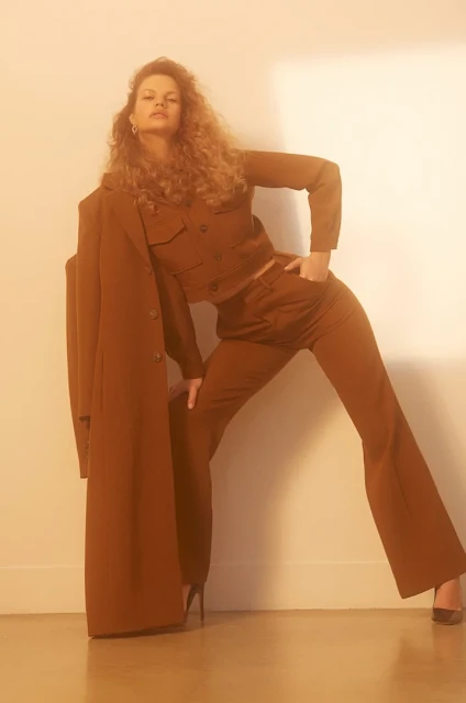 A woman poses in a brown two-piece outfit and heels, leaning against a white wall with a long brown coat draped beside her.
