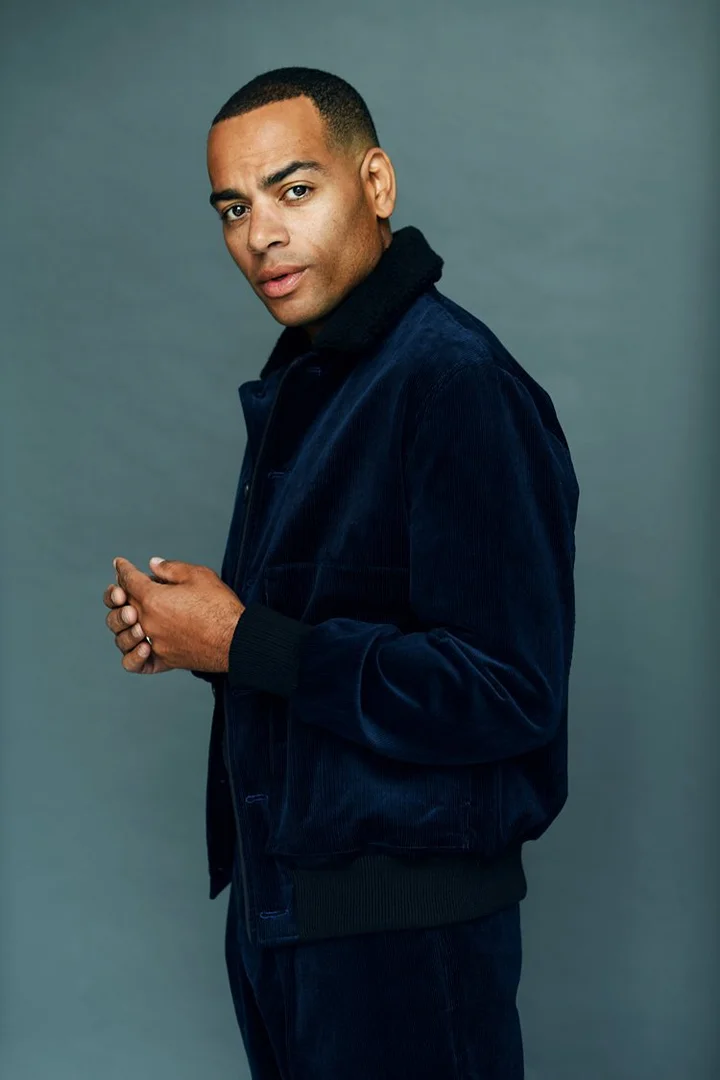 Close-up studio portrait of a dark-skinned adult male in a blue corduroy jacket, hands clasped, against a gray background.