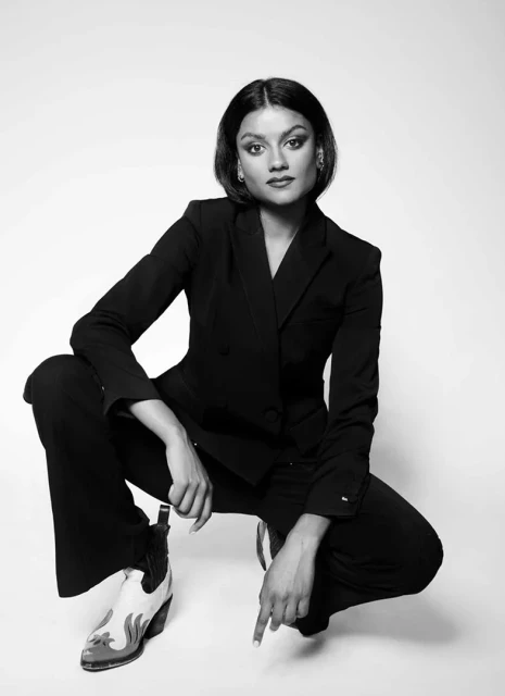 Elegant woman in a black blazer and pants poses crouching against a plain white backdrop, highlighted by her cowboy boot detail.