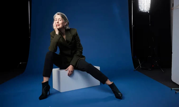 Older woman with grey hair wearing an olive blazer and black trousers seated on a white block against a blue backdrop