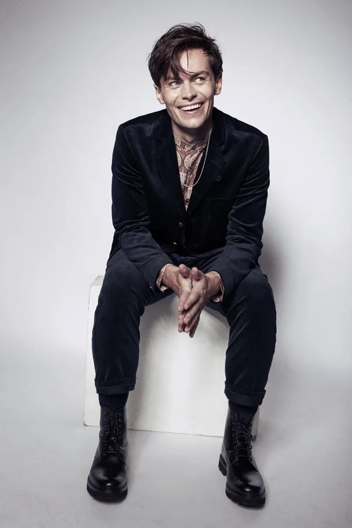 Smiling adult man sitting on white box in a photo studio wearing a velvet suit and boots with layered necklaces. 