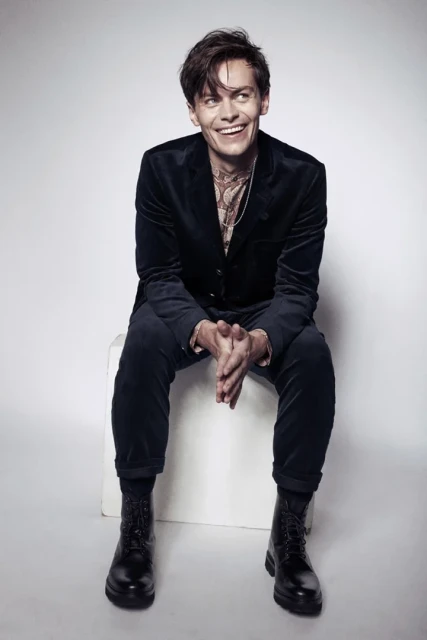 Smiling adult man sitting on white box in a photo studio wearing a velvet suit and boots with layered necklaces. 