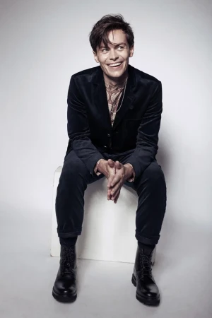 Smiling adult man sitting on white box in a photo studio wearing a velvet suit and boots with layered necklaces. 