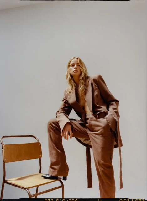 A fashionable blonde woman in a brown suit leans on a wooden chair against a white background, looking directly at the camera.