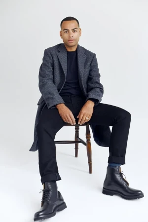Seated man wearing a grey tweed coat, black trousers, and leather boots against a white background