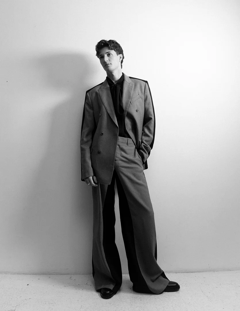 Young adult in a grey suit with black accents, standing against a white wall in a studio, looking at the camera with a neutral expression.