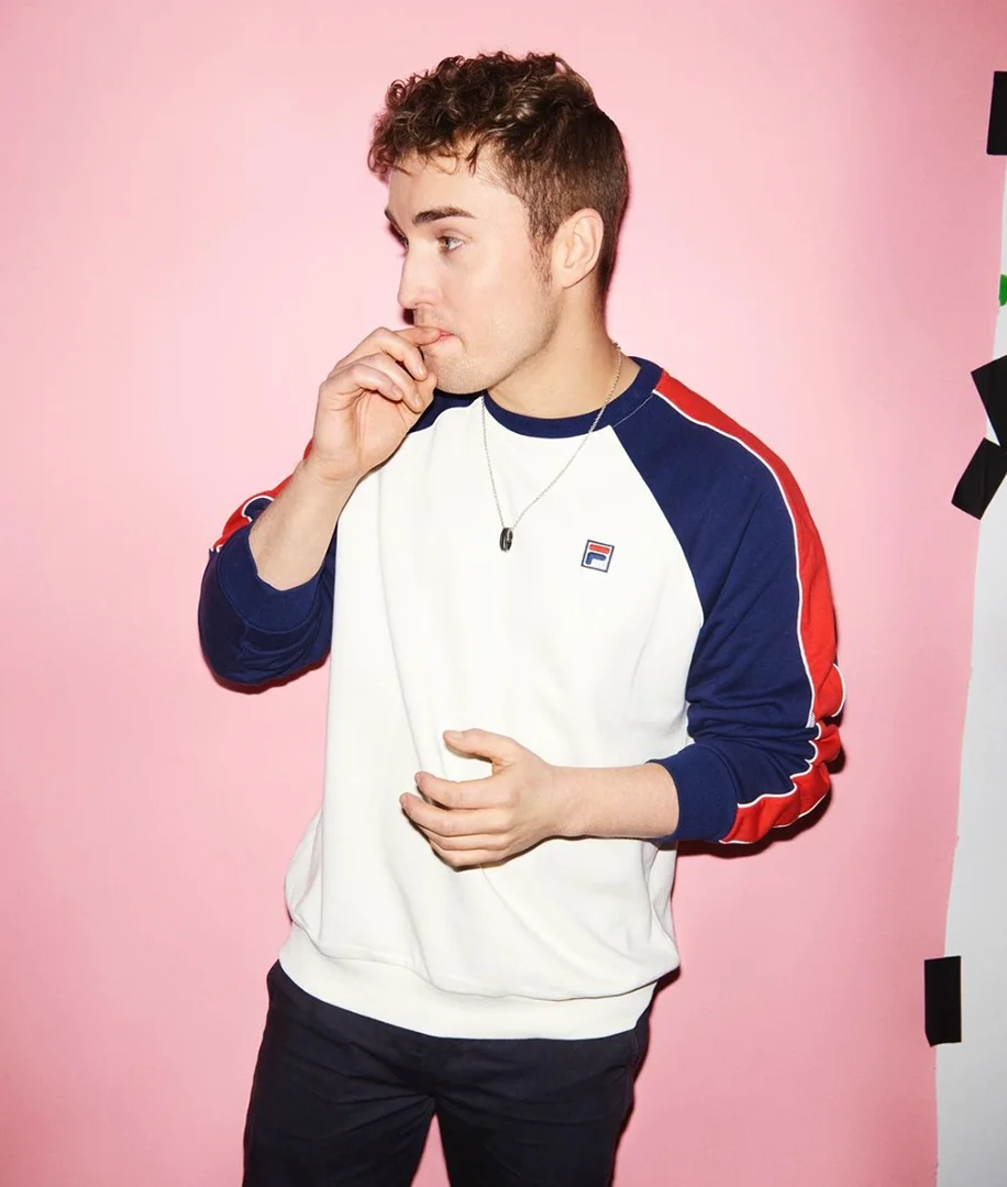 Portrait of a young man with curly brown hair in a white and blue sweatshirt against a pink background in a photo studio.