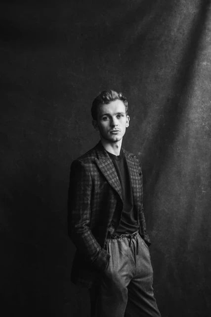 Monochrome portrait of a young man in a patterned blazer and casual pants posing indoors with hands in pockets against a dark backdrop.