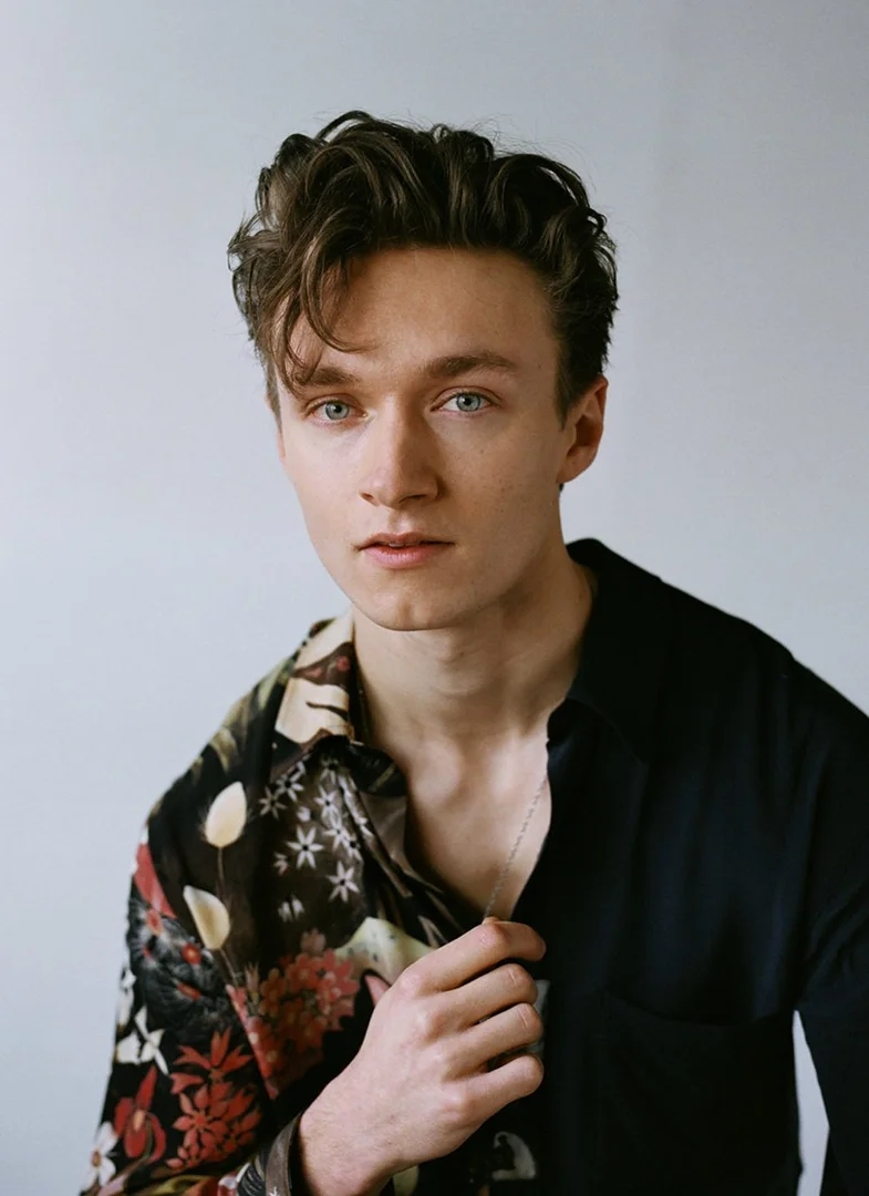 Portrait of a young adult male with dark wavy hair, wearing a floral shirt and looking directly at the camera against a plain backdrop.