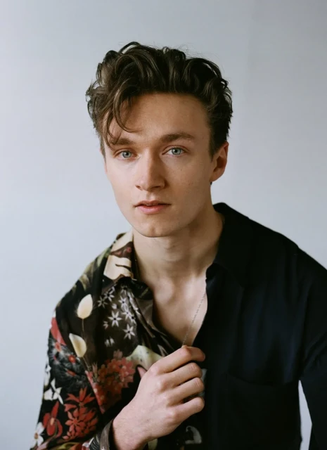 Portrait of a young adult male with dark wavy hair, wearing a floral shirt and looking directly at the camera against a plain backdrop.