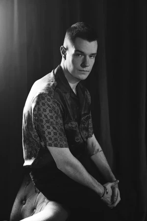 Monochrome portrait of a young man with short hair wearing a patterned short-sleeve shirt, seated against dark drapes.