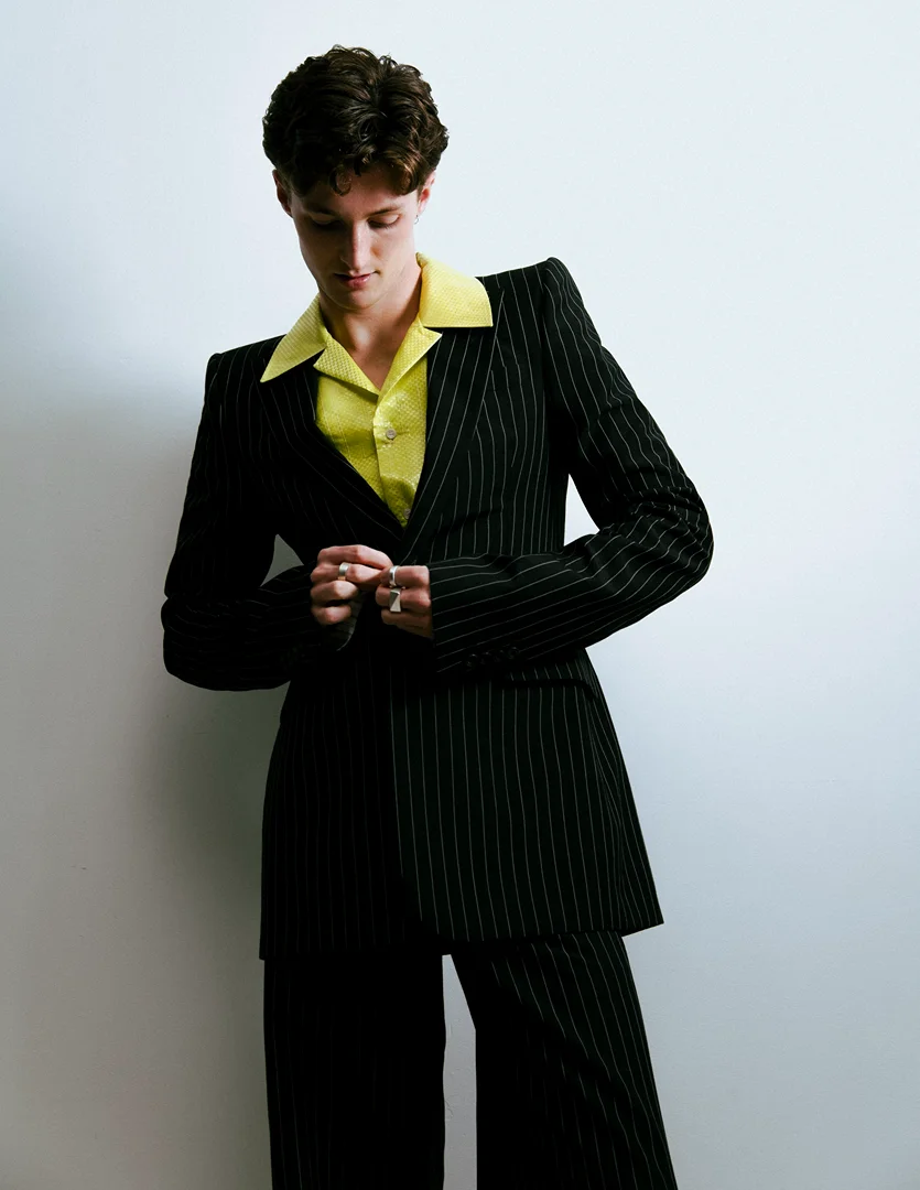 Young adult in a pinstripe suit fastens the jacket button, posing indoors with a bright yellow collared shirt.