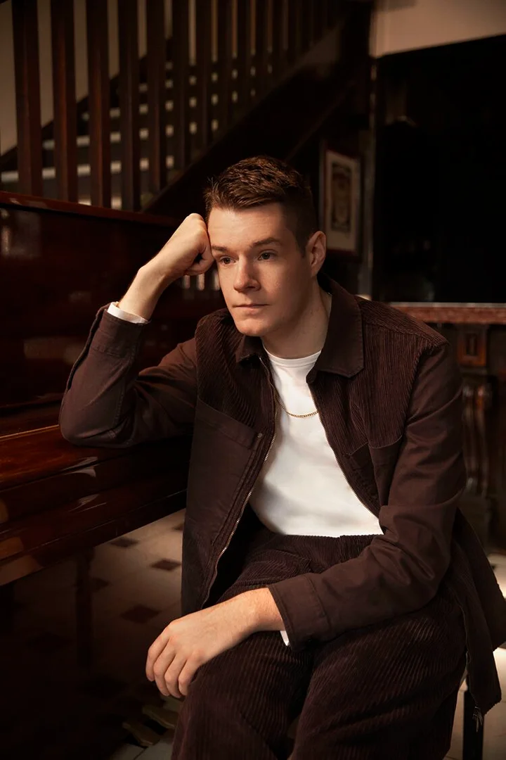 Portrait of a young adult male with short hair, wearing a brown corduroy jacket and white t-shirt, indoors with wooden background.