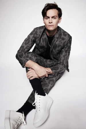 A man wearing a grey patterned suit and white sneakers seated on a white studio background.