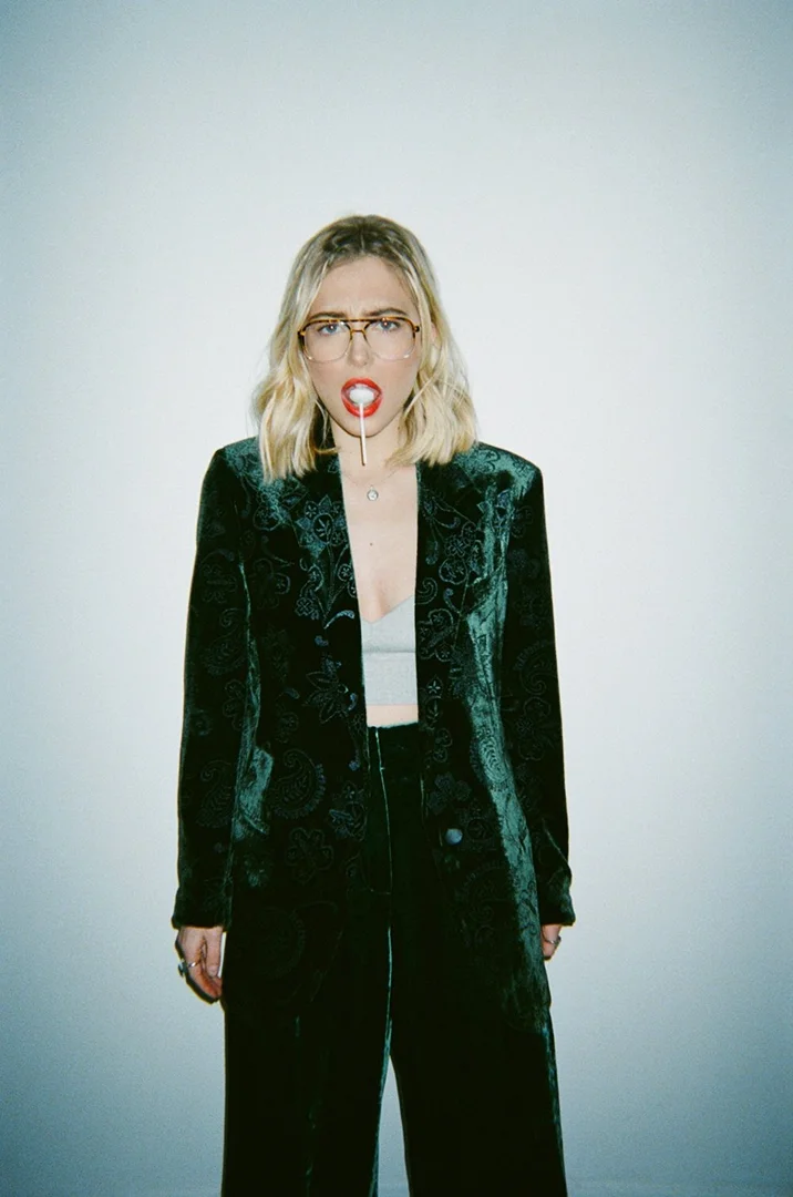 A woman with blonde hair, wearing glasses and a dark green velvet suit, poses indoors with a lollipop in her mouth against a white wall.