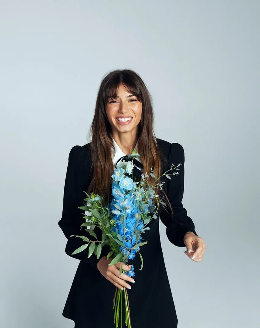 Smiling woman in a black dress holding a bouquet of blue flowers against a white background in a studio setting.