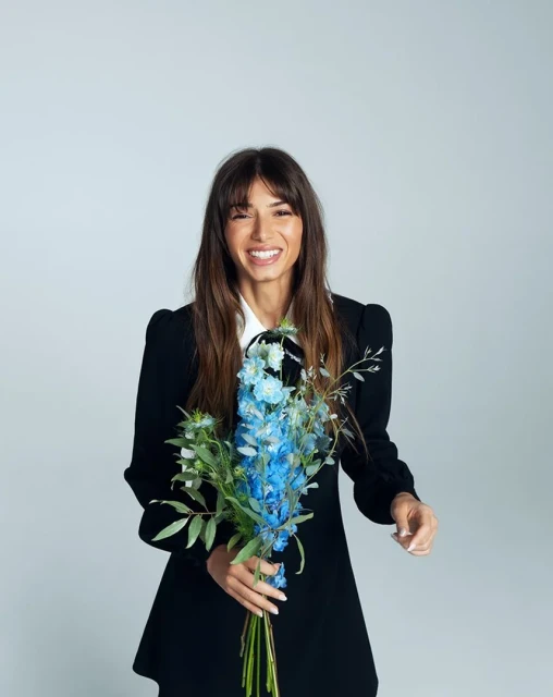 Smiling woman in a black dress holding a bouquet of blue flowers against a white background in a studio setting.