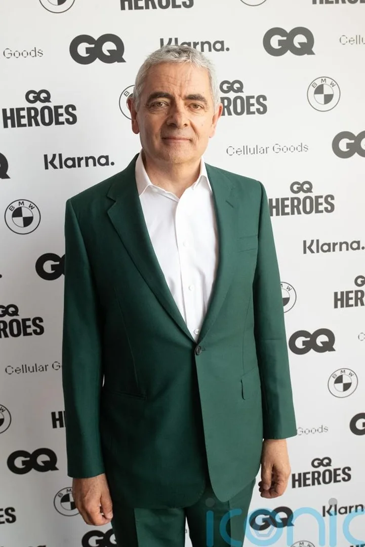 Rowan Atkinson poses in a green suit at GQ Heroes event against a branded backdrop, captured in a medium shot indoors.