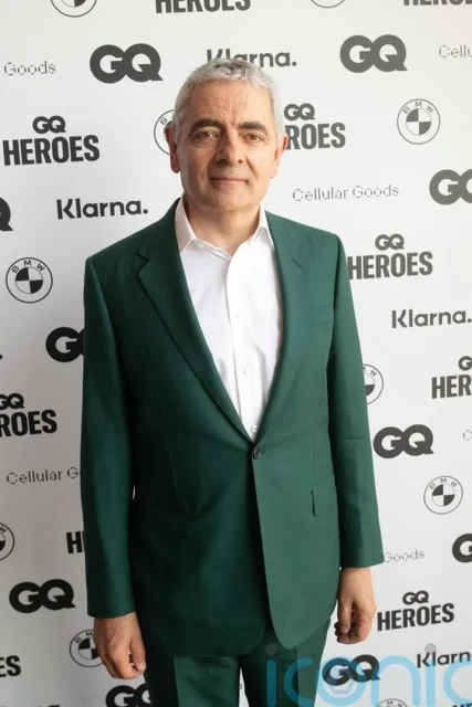 Rowan Atkinson poses in a green suit at GQ Heroes event against a branded backdrop, captured in a medium shot indoors.