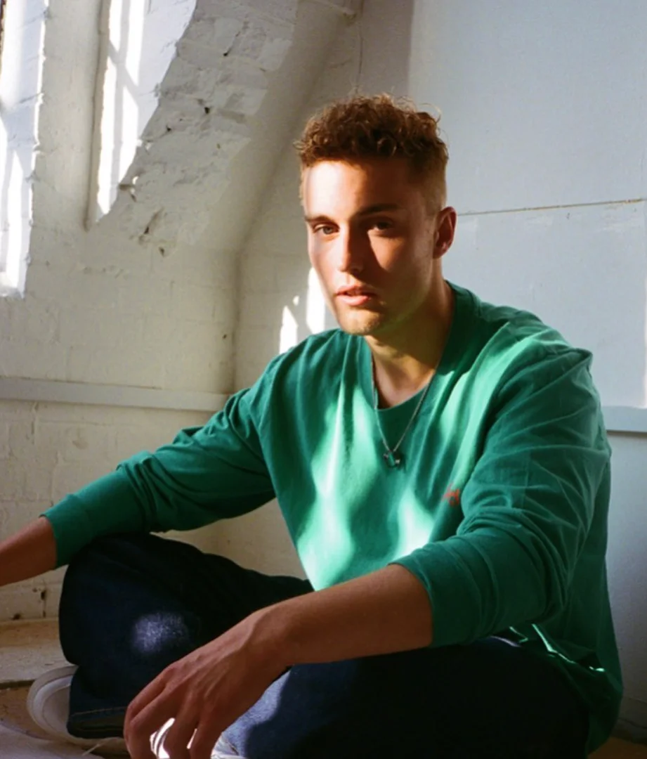 Young adult male with short blond hair poses in a bright indoor space wearing a green long-sleeved shirt and dark denim jeans.