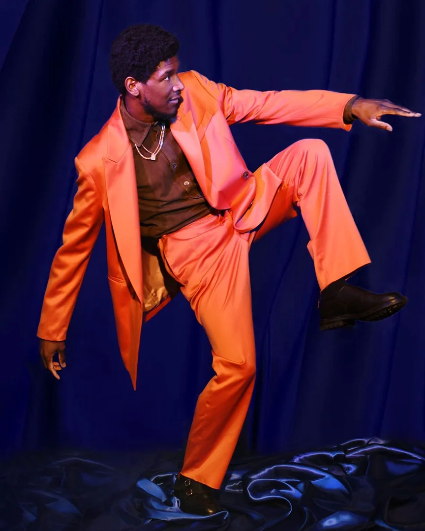 A Black male model poses in an orange suit against a dark blue draped backdrop with expressive movement, showcasing fashion and style.