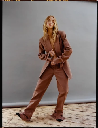 Model with long blonde hair wearing a brown pantsuit standing on a wooden floor against a grey backdrop