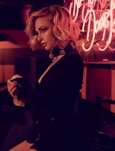A woman with blonde hair and dark clothing poses in front of a neon sign in a dimly lit indoor setting, highlighting fashion and portraiture.