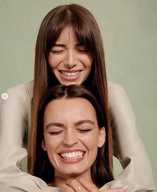 Two smiling adult women pose indoors with a pale green backdrop, one slightly behind the other. Both wear light-colored tops.