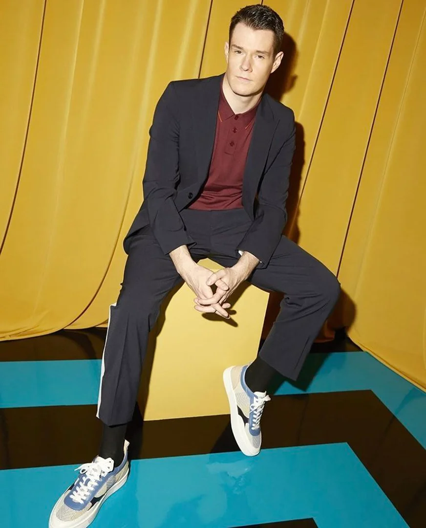Man in suit and sneakers sits against a golden backdrop. Dark suit with maroon polo shirt, sneakers with blue floor reflection.