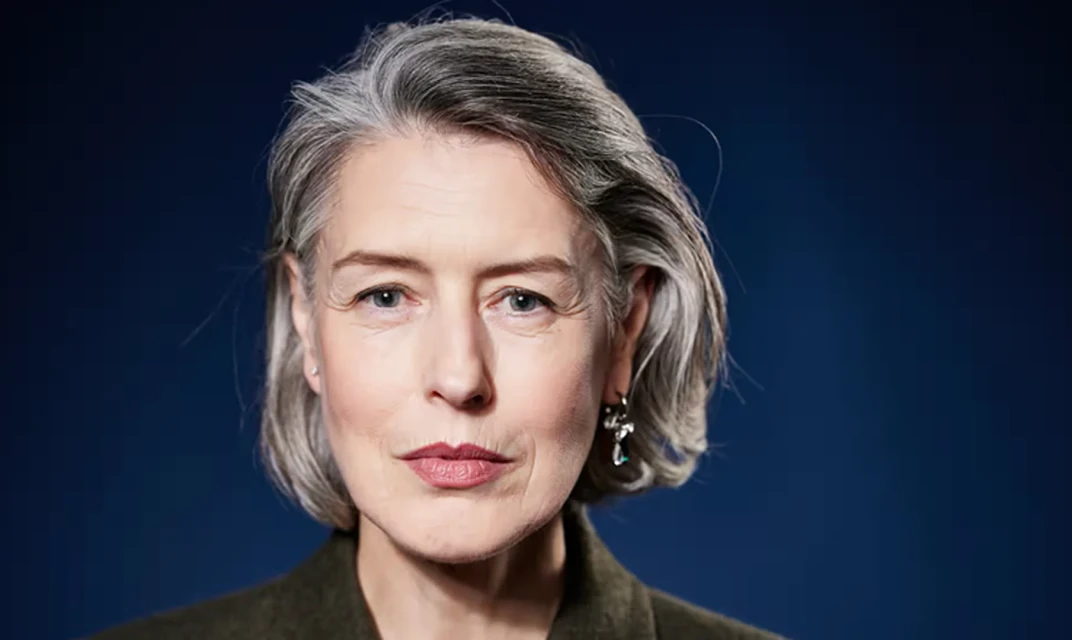 Portrait of a mature woman with grey hair and an olive-green jacket against a navy blue background. She is looking directly at the camera.