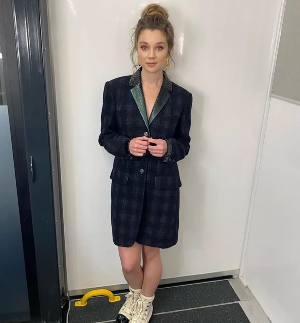Elegant woman in a plaid blazer dress, striking a pose indoors against a white wall, complemented by statement earrings and sneakers.