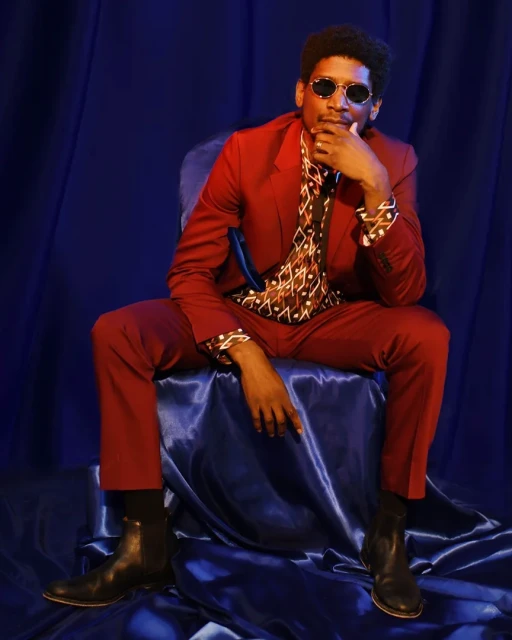 Portrait of stylish adult male wearing a burgundy suit, patterned shirt, and sunglasses sitting in front of a dark blue background.
