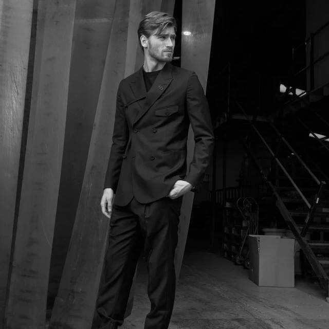 Black-and-white portrait of an adult male model posing indoors in a double-breasted suit, standing in front of a metal structure and stairs.