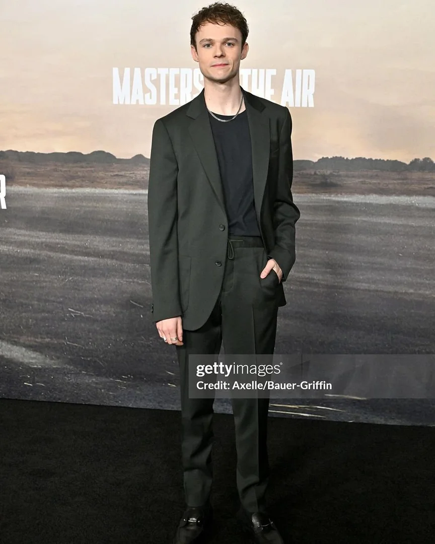 Actor stands on a red carpet wearing a dark suit in front of a 'Masters of the Air' promotional backdrop at a premiere event.