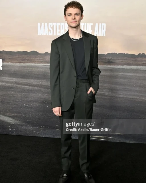 Actor stands on a red carpet wearing a dark suit in front of a 'Masters of the Air' promotional backdrop at a premiere event.