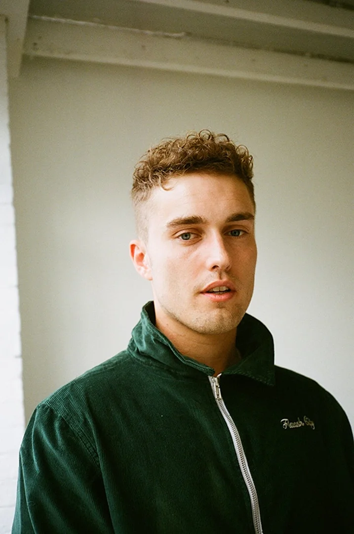 Young man with curly hair, wearing a corduroy jacket, poses indoors against a neutral background. Portrait shot of an attractive male.