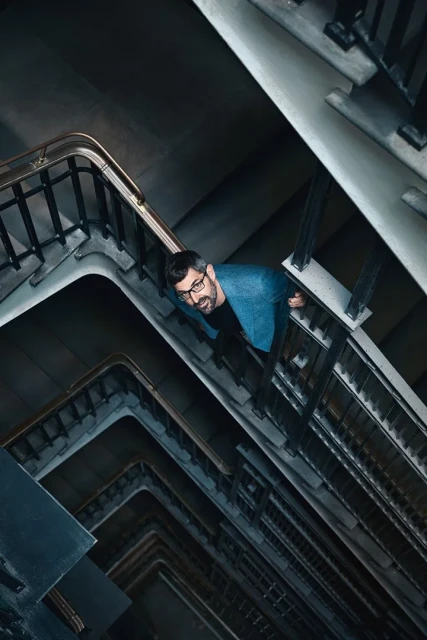 Low-angle shot of a man in glasses and a blue jacket standing in a concrete stairwell with metal railings, creating a geometric pattern.