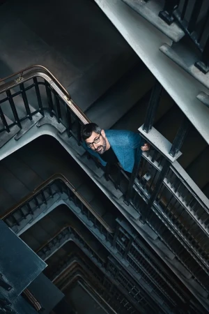 Low-angle shot of a man in glasses and a blue jacket standing in a concrete stairwell with metal railings, creating a geometric pattern.