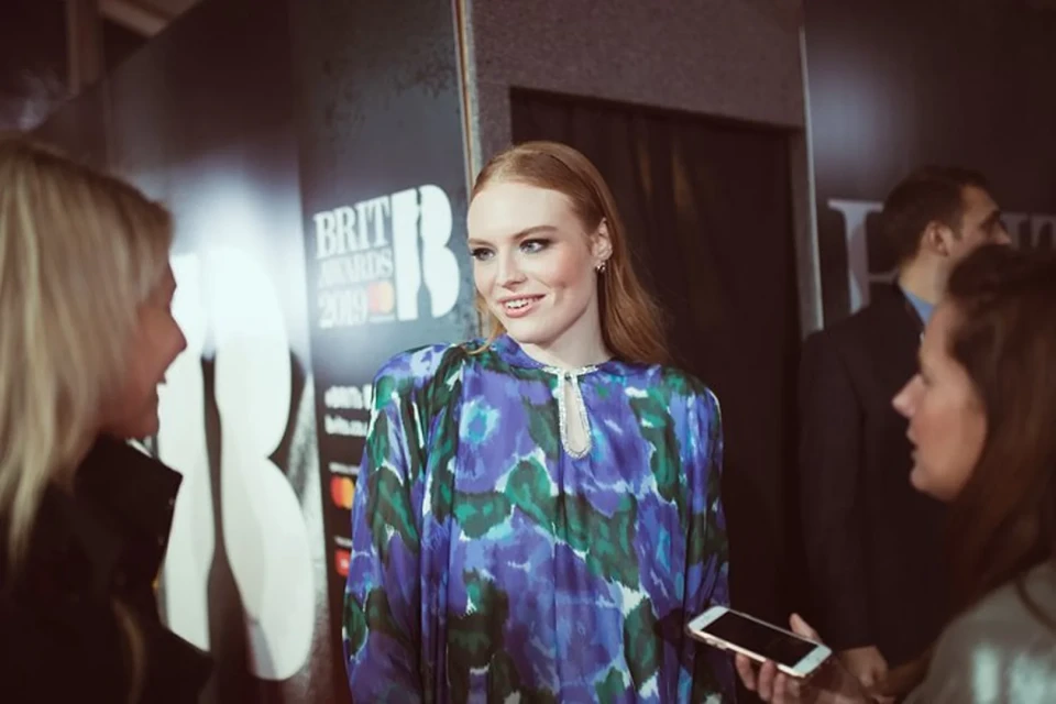 Red-haired woman in patterned dress photographed on a red carpet surrounded by event attendees and branding at an awards ceremony.