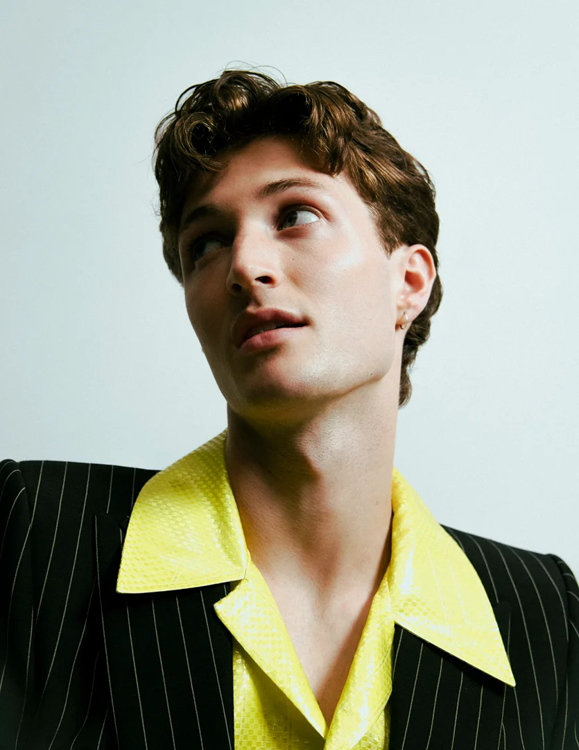 Portrait of a young man with brown curly hair wearing a pinstripe blazer and bright yellow collared shirt against a white background.