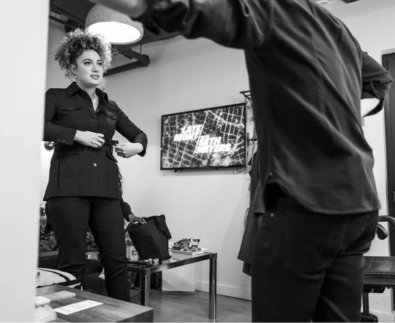 A woman in a black jacket adjusts her belt indoors, with a muted television in the background, in a monochrome photograph.