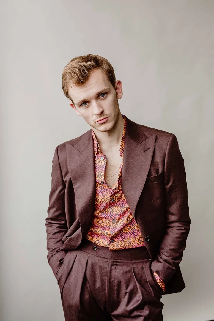 Portrait of a young adult male wearing a maroon suit jacket and pants with a colorful patterned shirt standing against a plain background.