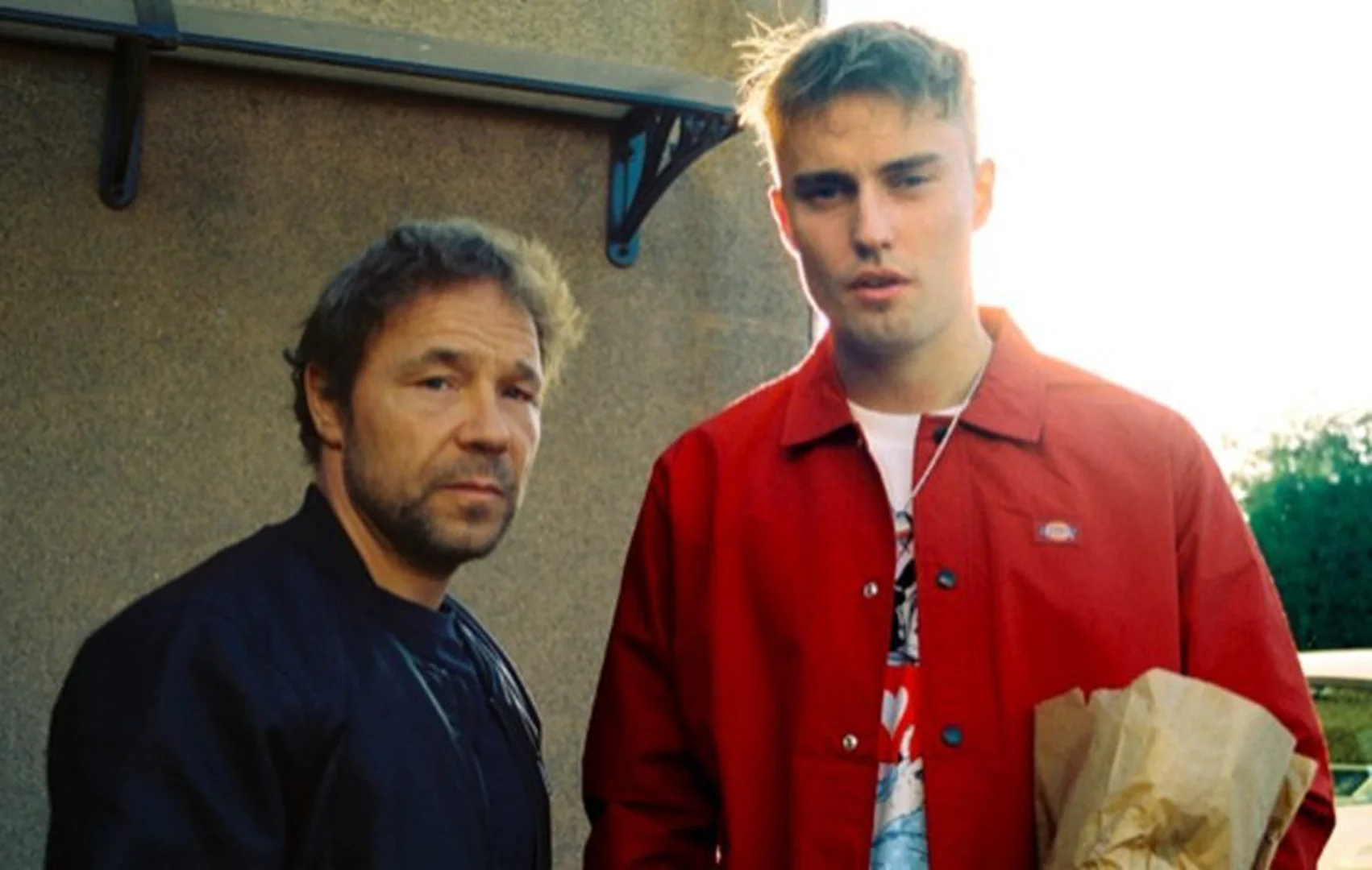 Two men stand outdoors in front of a building, one wearing a red jacket, with natural light and a shallow depth of field.