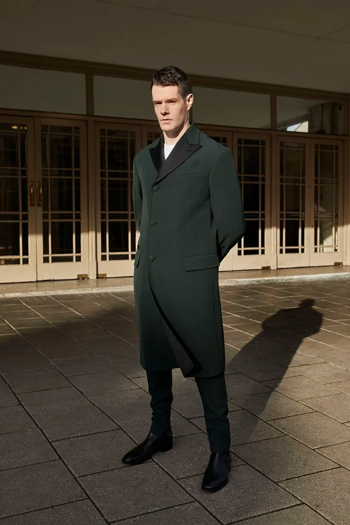 A stylish adult male stands outdoors on a stone plaza wearing a long, dark green coat, black pants, and black leather boots, looking at the camera.