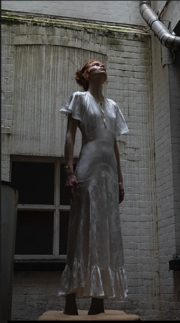 Woman in vintage dress looks up while standing in a courtyard with a brick wall background and metal pipes along the building.