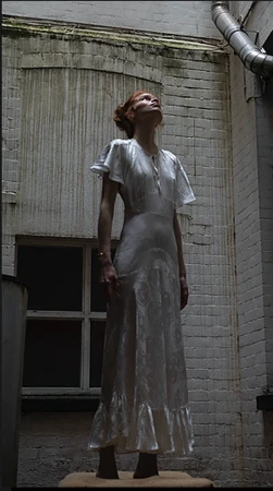 Woman in vintage dress looks up while standing in a courtyard with a brick wall background and metal pipes along the building.