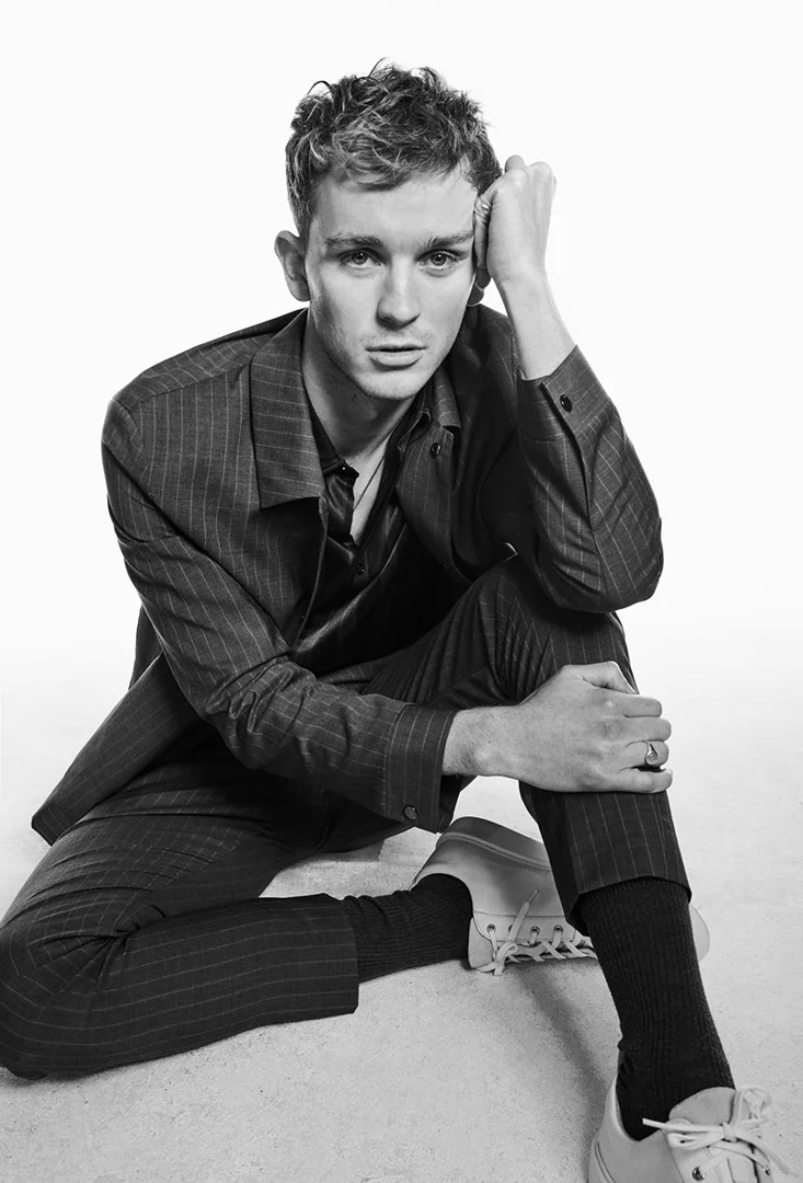 Black and white portrait of a young adult male with short hair, wearing a pinstripe suit and sneakers, sitting against a plain white background.