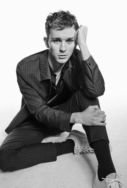 Black and white portrait of a young adult male with short hair, wearing a pinstripe suit and sneakers, sitting against a plain white background.