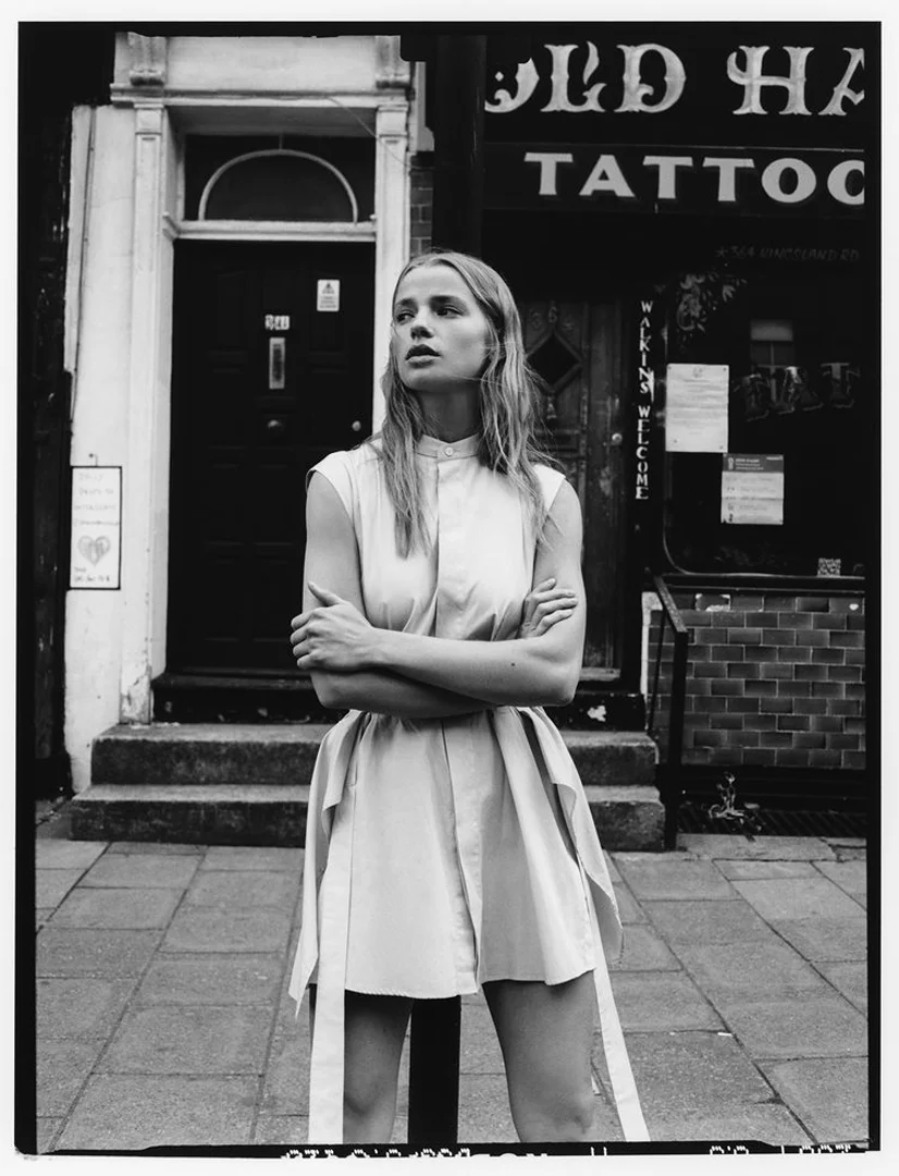 A young woman with blonde hair poses in a shirt dress in front of a tattoo parlor on a city street in a black and white editorial fashion photograph.