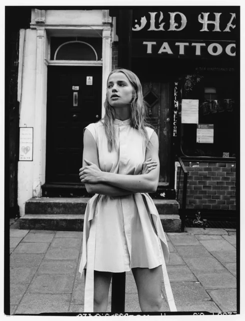 A young woman with blonde hair poses in a shirt dress in front of a tattoo parlor on a city street in a black and white editorial fashion photograph.